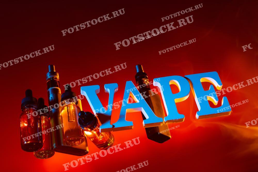 Фотография изображает электронные сигареты и флаконы с жидкостью для вейпинга, расположенные на фоне надписи "VAPE".